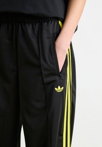 Black athletic pants with yellow stripes, elastic waistband, side pockets, and a small embroidered logo on the thigh. Smooth fabric texture.