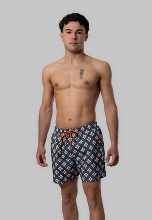 Giovane uomo senza maglietta con capelli neri ricci che indossa pantaloncini da bagno colorati e a fantasia, in piedi davanti a uno sfondo grigio uniforme.