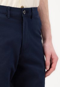Temně námořnické kalhoty s knoflíkovým zapínáním, bočními kapsami a poutek na opasek. Materiál má hladkou texturu s mírným leskem.