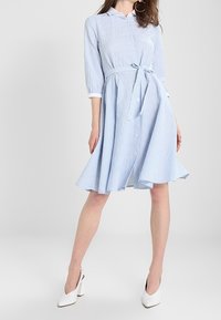 Kvinna iklädd en blå och vit rutmönstrad midiklänning med trekvartslånga ärmar och vita klackskor, stående med ett ben framåt.