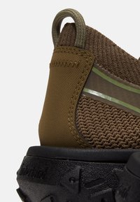 Primo piano del tallone di una sneaker marrone testurizzata con linguetta per tirare, tomaia a coste, striscia riflettente e suola in gomma nera robusta.