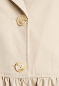 Beżowa sukienka z materiału z kołnierzem klapowym, z dwoma dużymi, kremowymi guzikami i marszczonym falbaną na dole. Gładka faktura.