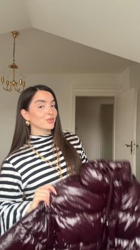 Tânără cu păr lung și închis la culoare, purtând o cămașă cu dungi și bijuterii aurii, ține o jachetă strălucitoare de culoare mov închis într-o cameră cu nuanțe neutre.