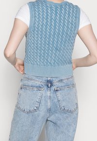 Gilet en tricot bleu clair avec un motif en relief, ourlet en grosses côtes et design sans manches, associé à un jean bleu clair taille haute.