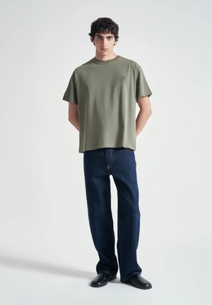 Giovane uomo in piedi con le mani dietro la schiena, indossa una maglietta verde oliva, jeans blu scuro e scarpe nere su uno sfondo semplice.