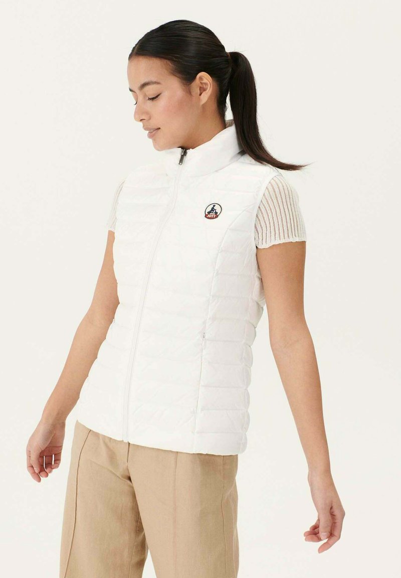 Chaleco acolchado blanco con cuello alto, cierre de cremallera y parche con el logo en el lado izquierdo del pecho, combinado con una camiseta de manga corta con canalé.