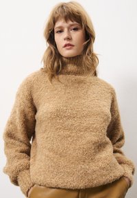 Beige, fuzzy gebreide trui met een hoge col en lange mouwen. Zachte textuur en ruime pasvorm, gecombineerd met hooggewaiste bruine broek.