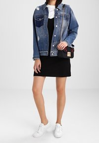 Nainen, jolla on päällään kulunut farkkutakki mustan mekon ja valkoisten tennareiden kanssa, pitää kädessään pientä mustaa olkalaukkua, jossa on kultainen solki.