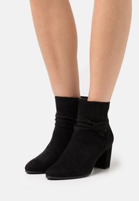 Bottines en daim noir avec talon évasé, bout pointu et fronces décoratives avec un nœud au niveau de la cheville. Texture lisse.