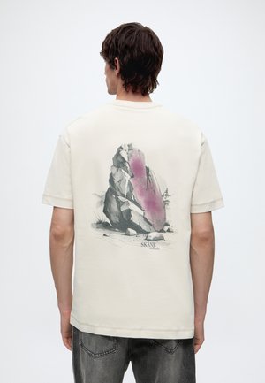 Man met de rug naar de camera in een wit T-shirt met een grote rotsafbeelding en tekst "Skåne wetlands" op de achterkant.