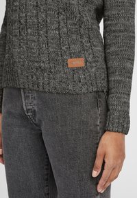 Gros plan d'une personne portant un pull en maille torsadée gris foncé avec une étiquette de marque marron et un jean en denim gris foncé, main reposant à côté.