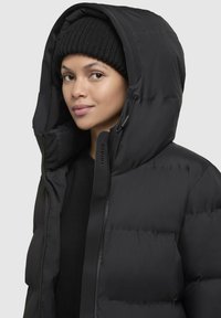 Femeie purtând o pălărie neagră din tricot și un hanorac pufos negru cu glugă, privind înainte cu o expresie neutră pe un fundal simplu.