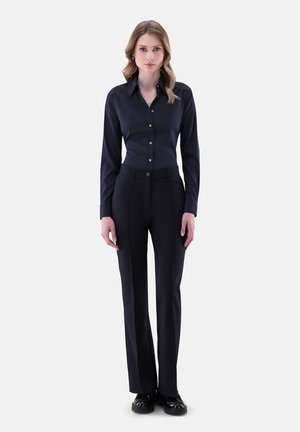 Donna in piedi di fronte, che indossa una camicia a bottone a pois blu scuro, pantaloni neri e scarpe nere su uno sfondo neutro.