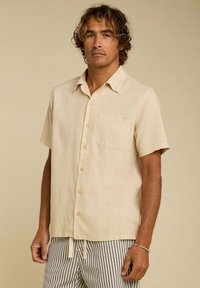 Chemise à manches courtes beige clair avec un bouton sur le devant et une poche poitrine, fabriquée dans un tissu texturé, comportant une étiquette logo discrète et une coupe décontractée.