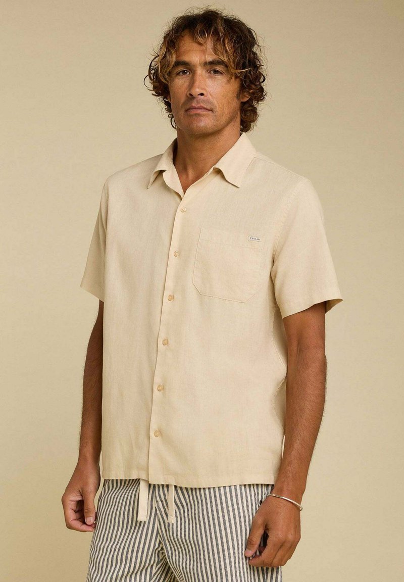 Chemise à manches courtes beige clair avec un bouton sur le devant et une poche poitrine, fabriquée dans un tissu texturé, comportant une étiquette logo discrète et une coupe décontractée.