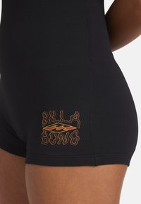 Czarny kostium kąpielowy z teksturowanej tkaniny. Posiada wyszywane na biodrze logo "Billabong" w kolorze żółtym i czerwonym, przedstawiające wzór fali.