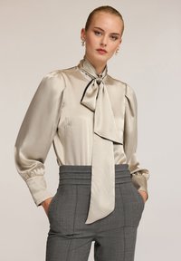 Blusa satinata beige con colletto a fiocco, maniche lunghe a sbuffo e dettagli scuri, abbinata a pantaloni grigi sartoriali a vita alta.