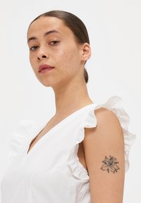 Bjela majica bez rukava s nabranim rubovima, v-izrezom i mekanom teksturom. Model ima cvjetni tattoo na gornjem dijelu ruke.