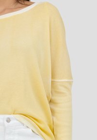 Vaaleankeltainen ribbed pitkähihainen paita, jossa on leveä kaula-aukko ja valkoiset reunat. Pehmeä rakenne, rento istuvuus ja näkyvät ompeluyksityiskohdat hihoissa.