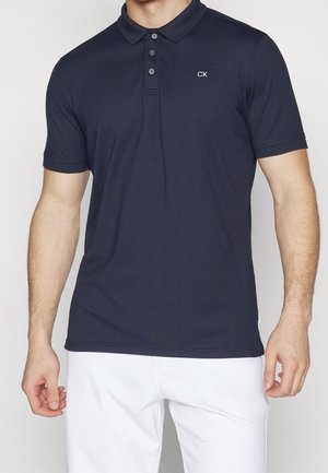Man in een marineblauw polo met korte mouwen met drie knopen en wit "CK" logo, gecombineerd met witte broek.