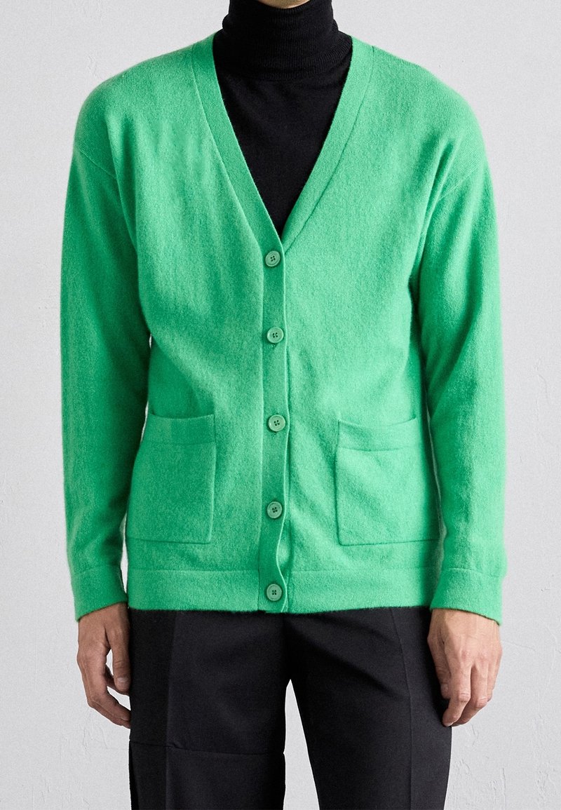 Grüne Strickjacke mit Knöpfen und Fronttaschen, getragen über einem schwarzen Rollkragenpullover von einer Person, die vor einem hellen Hintergrund steht.