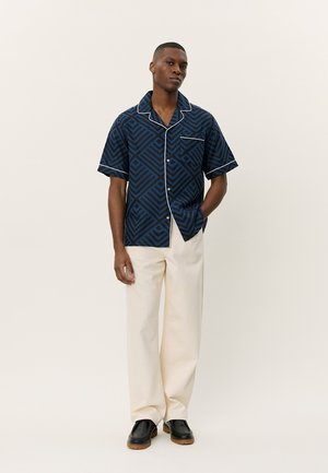 Marineblauwe kortekousen HEM met geometrisch patroon en witte bies, gecombineerd met crème broek en zwarte schoenen, staande tegen een neutrale achtergrond.
