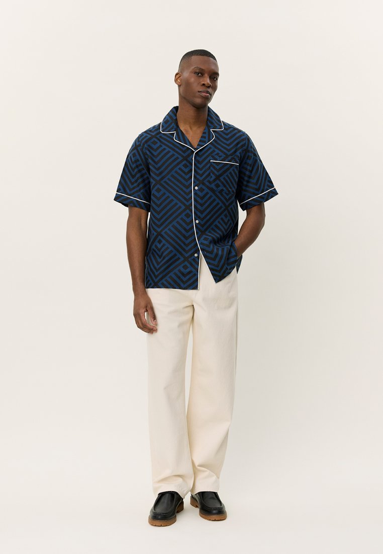 Camisa de manga curta azul-marinho com padrão geométrico e acabamento branco, combinada com calças creme e sapatos pretos, de pé contra um fundo neutro.