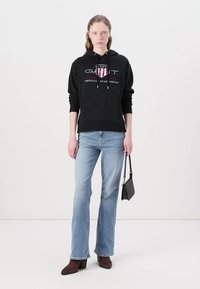 Black hoodie with white and red logo, paired with light blue flared jeans. Model carries a small black handbag and wears brown heeled boots.