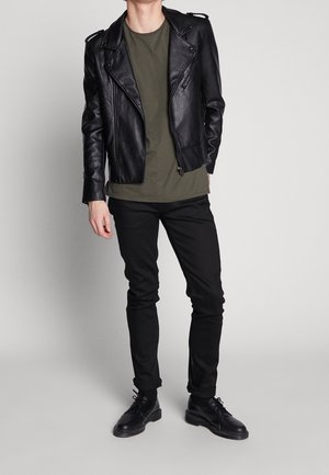 Chaqueta de motorista de cuero negro con cremalleras plateadas, camiseta verde, jeans ajustados negros y zapatos negros con cordones. Atuendo casual sobre un fondo liso.