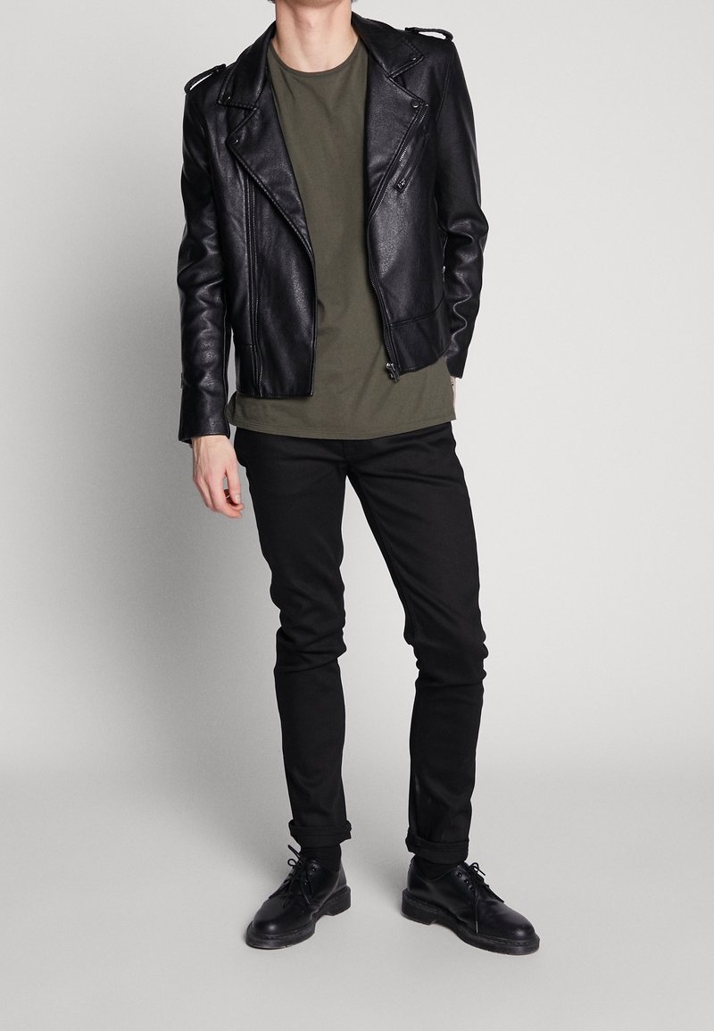 Sort læder bikerjakke med sølvfarvede lynlåse, grøn t-shirt, sorte skinny jeans og sorte snørebåndssko. Casual outfit på en ensfarvet baggrund.