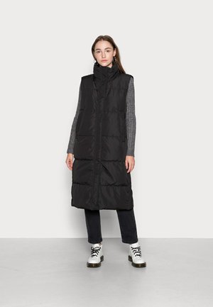 Gilet matelassé noir avec col haut, texture en piqué et fermeture éclair à l'avant ; porté sur une chemise grise à manches longues et associé à un pantalon sombre et des bottes blanches.