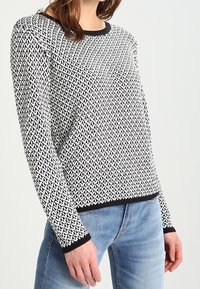 Femme portant un pull tricoté texturé noir et blanc avec un bord noir, associé à un jean bleu clair, debout devant un fond uni.