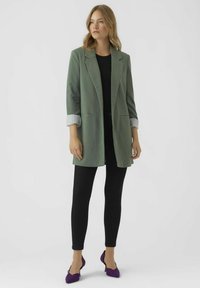 Světlě zelený dlouhý blazer se strukturovaným designem, se dvěma předními kapsami a ohrnutými pruhovanými rukávy. Skombinováno s černými legínami a fialovými botami.