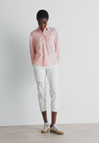 Camicia a maniche lunghe rosa chiaro con bottone frontale e taschino, abbinata a pantaloni bianchi a vita alta sfrangiati e sneakers grigio chiaro.