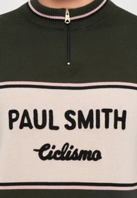 Maglione verde a maglia con colletto alto, zip dorata e testo nero in grassetto che dice "PAUL SMITH" sopra "Ciclismo" su uno sfondo chiaro.