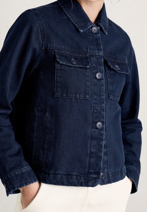 Donkerblauwe spijkerjas met knopen aan de voorkant en klepzakken, gedragen met handen in de zakken van een witte broek, bovenlichaam zichtbaar.