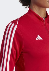Red athletic jacket featuring white stripes on sleeves, high collar, and zip closure. Made from smooth, textured fabric with a logo accent.
