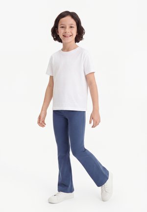 Kind mit dunklen Haaren lächelt, trägt ein weißes kurzärmeliges Hemd, blaue Hosen und weiße Turnschuhe, steht auf einem Bein vor weißem Hintergrund.