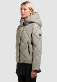 Quiltet puffer-jakke i dæmpet grå med hætte, med en sidelomme med lynlås og et mærke på venstre skulder. Blød, glat tekstur.