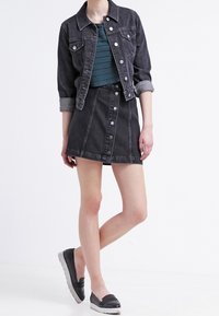 Kobieta ubrana w czarną dżinsową kurtkę i spódnicę z niebieską, teksturowaną górą oraz czarnymi butami na płaskiej podeszwie, stojąca na tle białym.