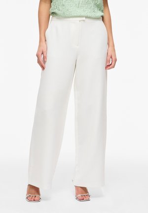 Femme portant un pantalon blanc taille haute à jambes larges avec poches latérales et talons argentés ouverts à l'avant, debout devant un fond blanc.