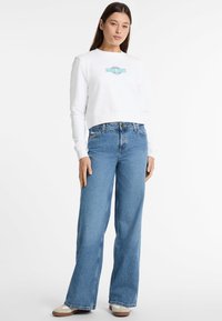 Valkoinen lyhyt sweatshirt, jossa on "Guess Jeans" -logo, yhdistettynä sinisiin leveälahkeisiin farkkuihin ja beigeihin lenkkareihin. Yksinkertainen muotoilu, rento istuvuus.