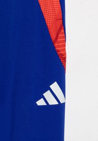 Blauer Sportstoff mit einem strukturierten roten Muster auf einer Seite, das ein auffälliges weißes Adidas-Logo in diagonalem Verlauf zeigt.