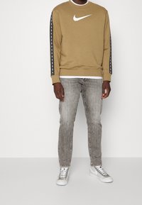 Sudadera de Nike en caqui con logo blanco, presenta acentos de rayas negras en las mangas. Combinada con jeans gris claro y zapatillas plateadas.