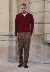 Czerwony sweter polo z teksturowanym splotem, w połączeniu z brązowymi spodniami w kratę i czarnymi loafersami. Strój charakteryzuje się luźnym krojem i klasycznym stylem.
