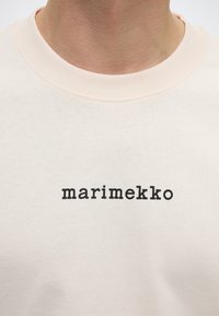 Κοντινή λήψη ενός μπεζ μπλουζιού με λαιμόκοψη, με τη λέξη "marimekko" τυπωμένη με μικρά μαύρα γράμματα στο στήθος.