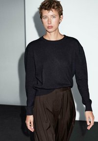 Pull en maille noir avec un col rond et des épaules tombantes, associé à un pantalon plissé marron foncé. Texture lisse, palette de couleurs neutres.