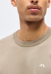 Κοντινό πλάνο ενός ατόμου που φοράει μπεζ πουλόβερ με στρογγυλή λαιμόκοψη και μικρό λευκό γεωμετρικό λογότυπο στο στήθος.