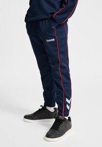 Mörkblå träningsbyxor med röda sidoframmar, med "Hummel"-logotypen och ett chevronmönster på det nedre benet, tillverkade av ett slätt tyg.