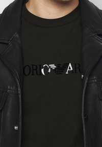 Primo piano di una persona che indossa una giacca di pelle nera sopra una maglietta nera con un logo e un emblema metallici sul petto.